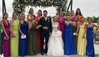 Camila e Giovanni com as madrinhas de vestidos coloridos diante do arco de flores